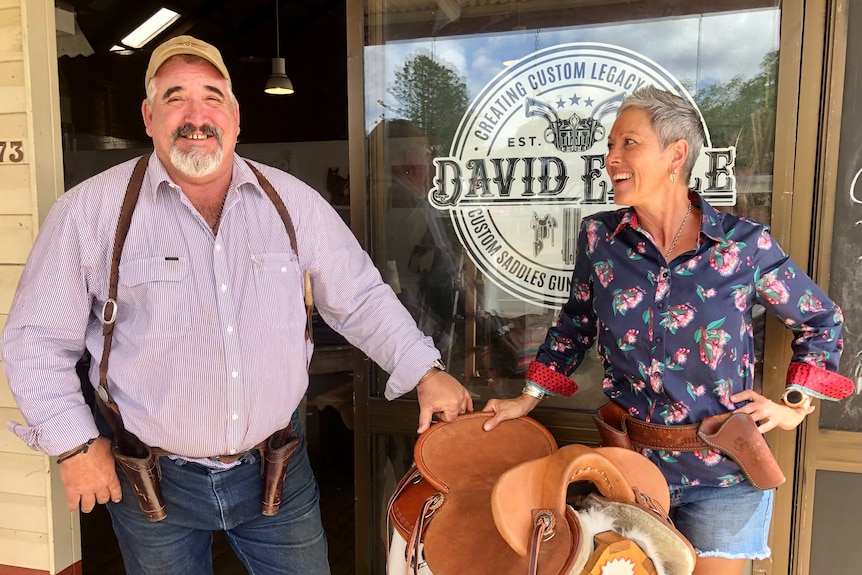 Two smiling people stand beside a saddle in front of their saddlery.