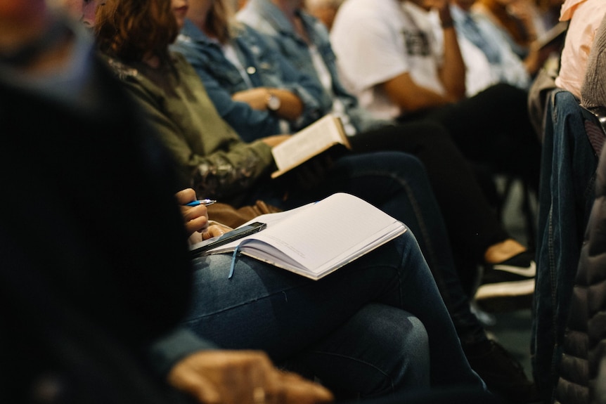 A photo of a line of people sitting with notebooks and pens in their laps.