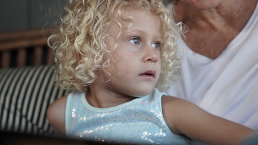 A young girl with striking blonde curls and blue eyes looks off camera.