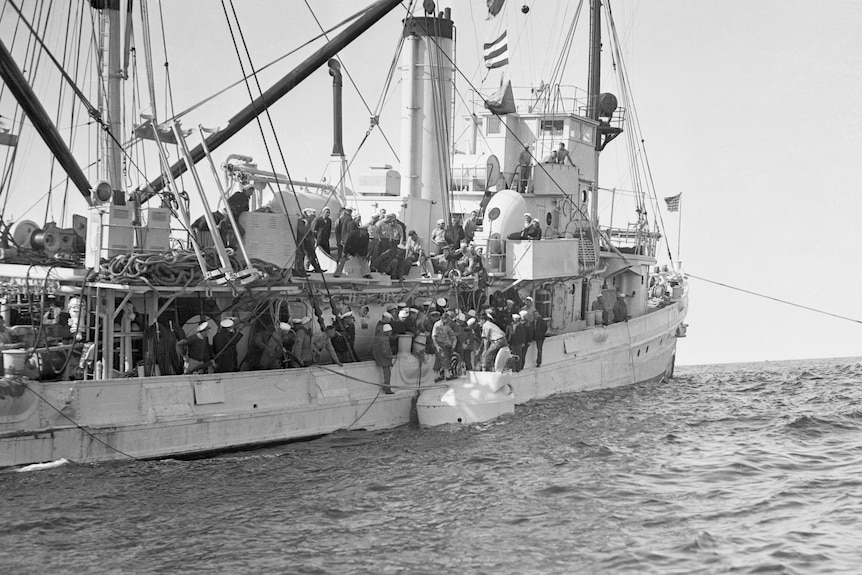 A small diving vessel hoisted up next to a large ship in the ocean. Divers and ship crew members are near the diving vessel