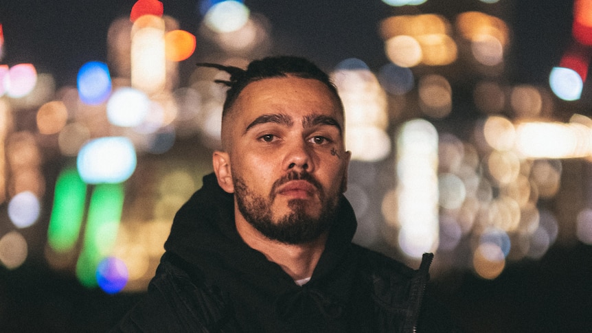 Kobie Dee stands in a hoodie against the blurry lights of a city skyline