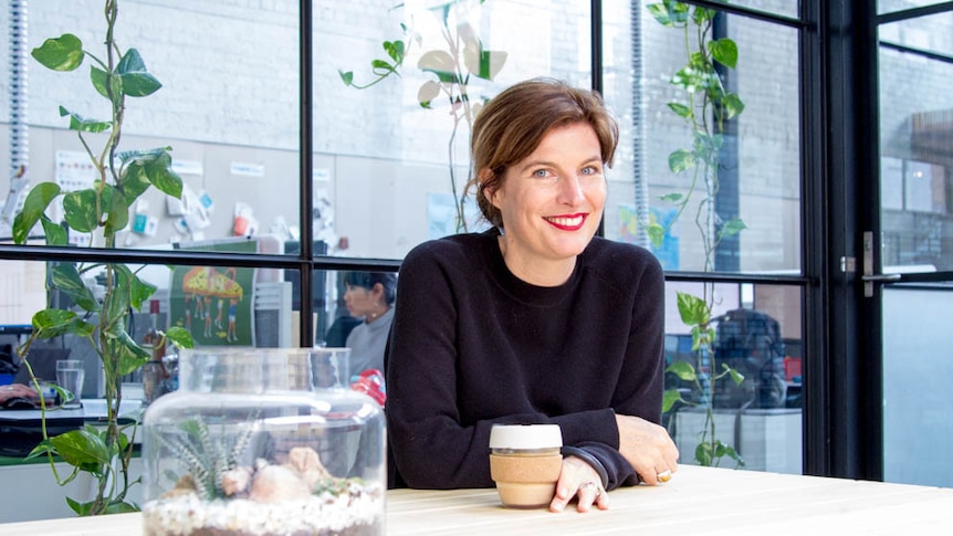 A woman smiles while sitting holding a glass coffee cup, open plan office through window in background.