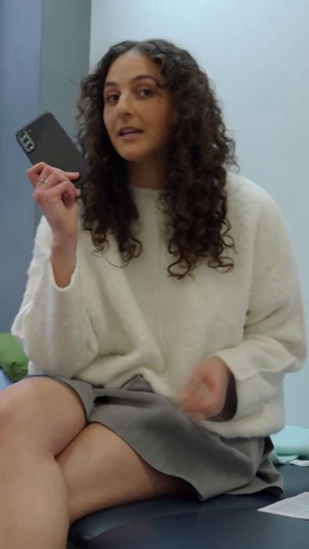 A young woman with medium-light-tone skin and curly hair holds a smartphone in her hand as she looks into the lens