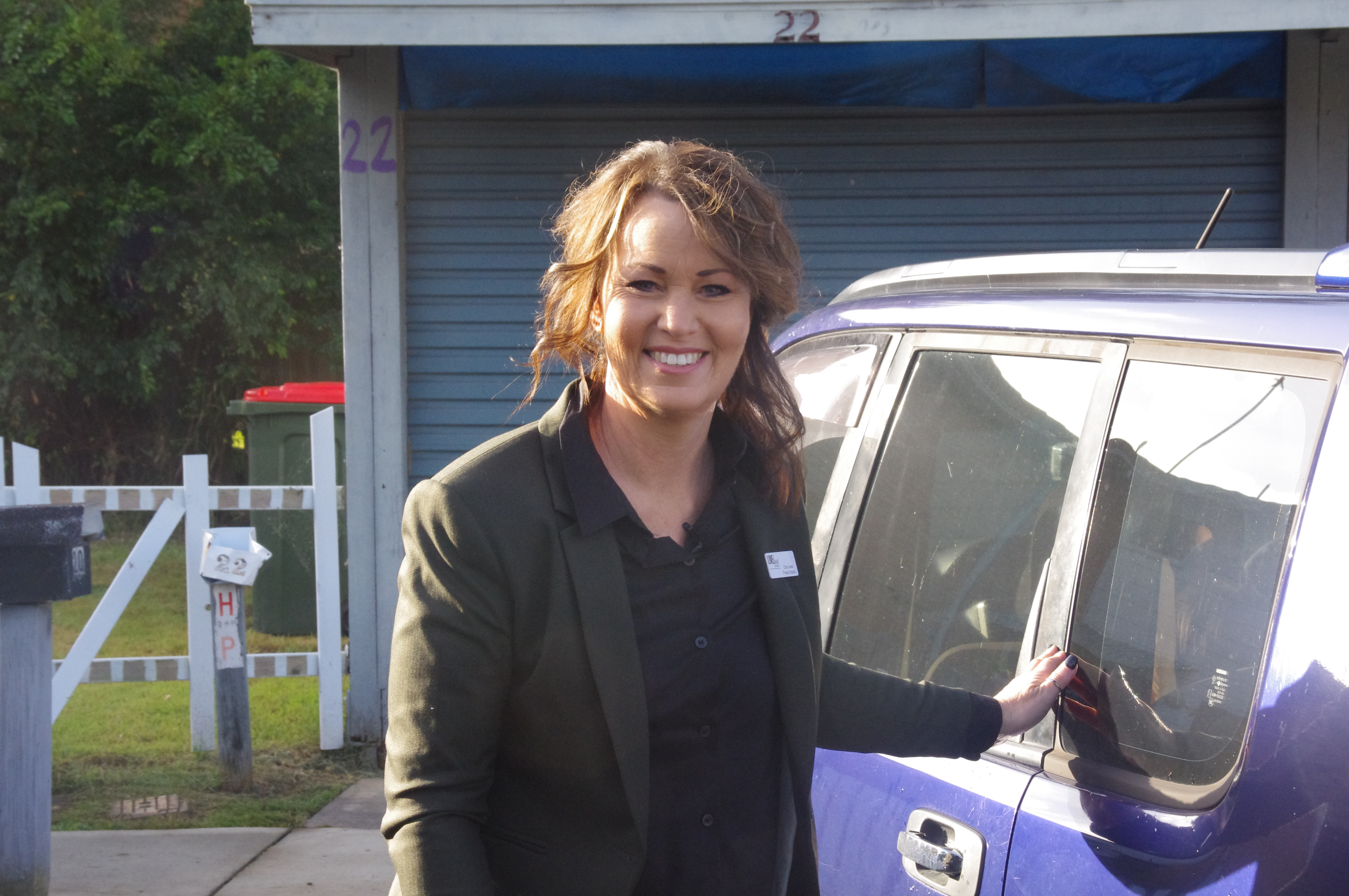A smiling woman stands in front of a blue car.