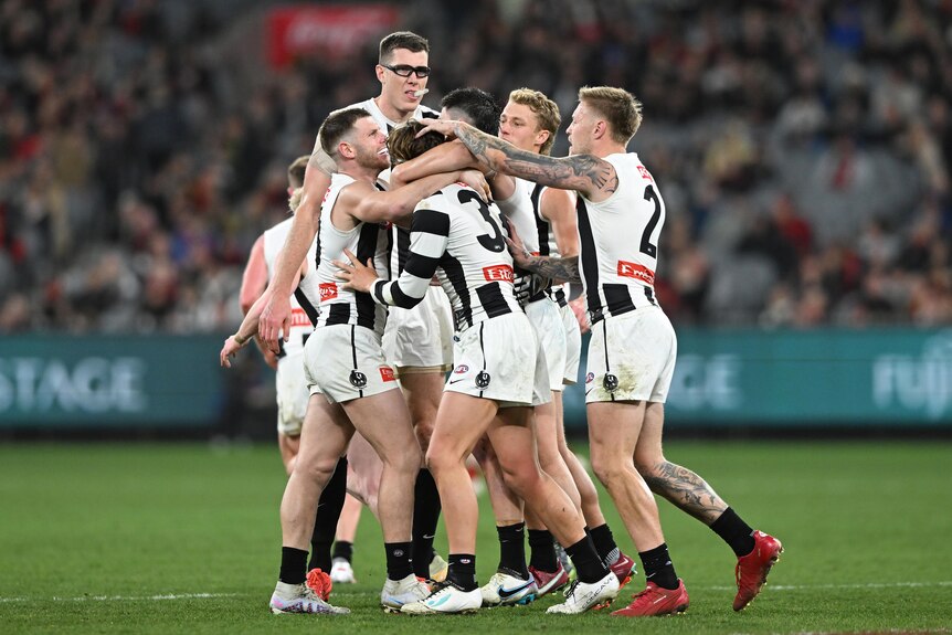 AFL players wearing black and white stripes converge in celebration in the middle of the field. 