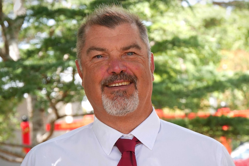 A man wearing a white shirt and red tie smiles and poses for a camera