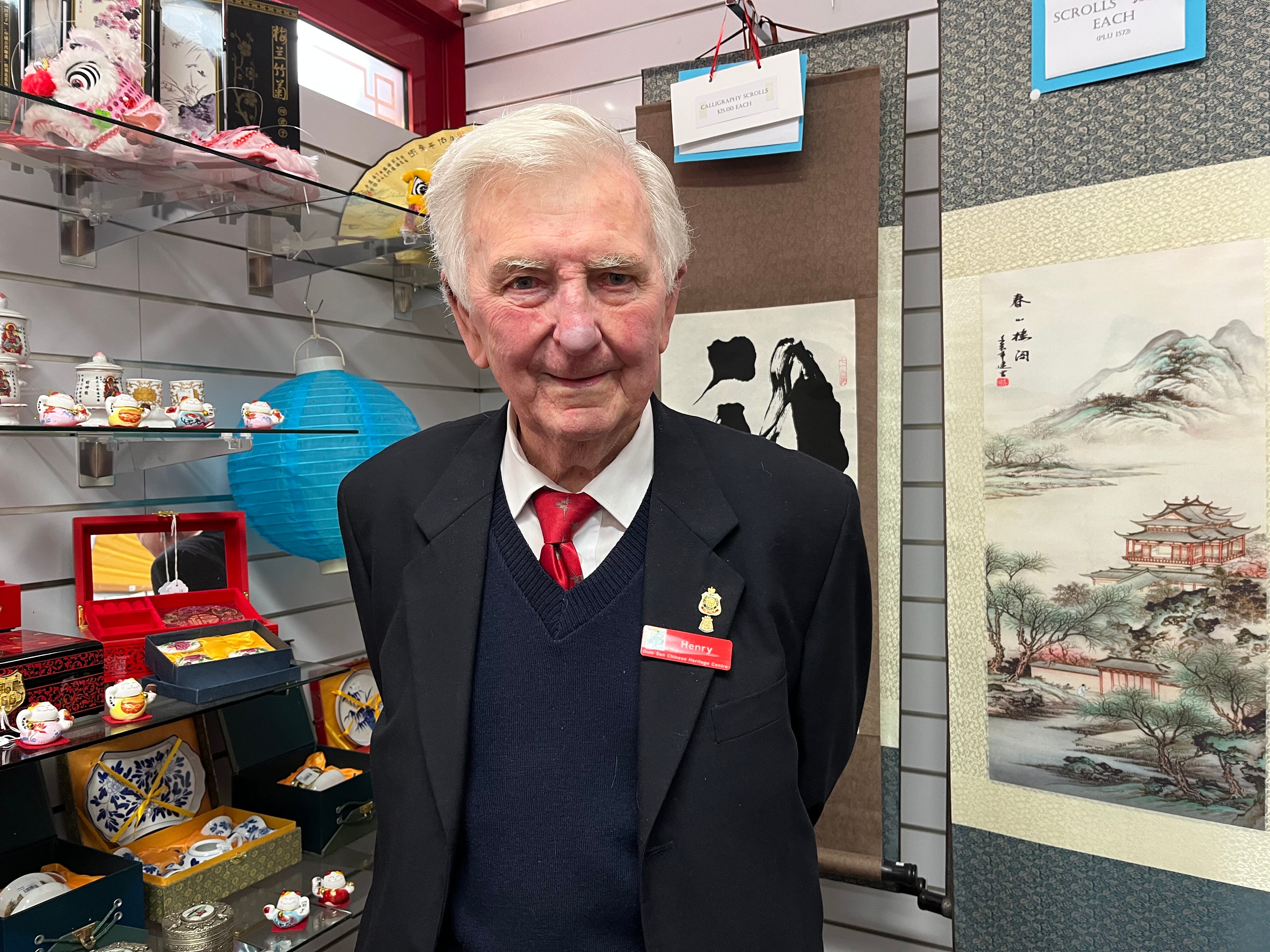 An older man with white hair wears a black blazer and red name tag over a navy sweater, white shirt and red tie gift shop behind