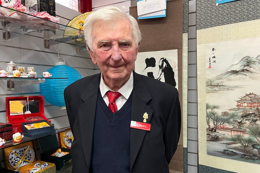 An older man with white hair wears a black blazer and red name tag over a navy sweater, white shirt and red tie gift shop behind