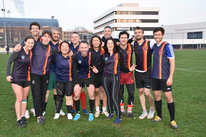 A group of 13 people dressed in the same uniforms stand together on a rugby field for a photo.