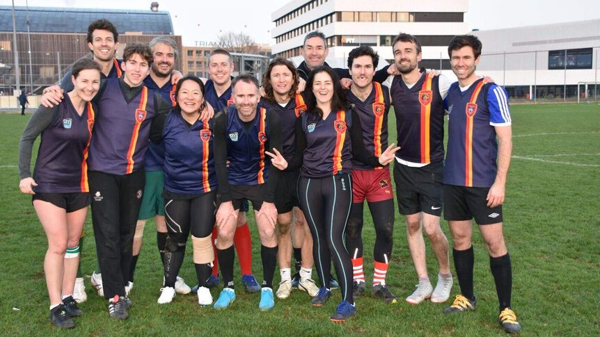 A group of 13 people dressed in the same uniforms stand together on a rugby field for a photo.
