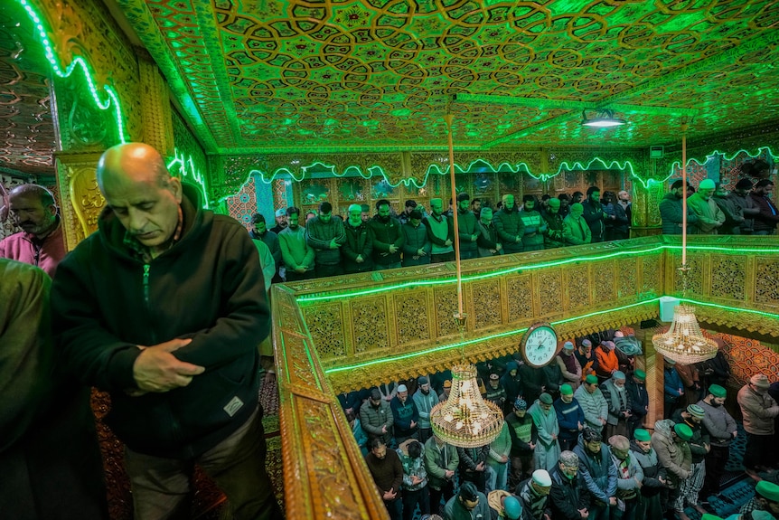 Worshipers pray inside a detailed mosque with green lighting