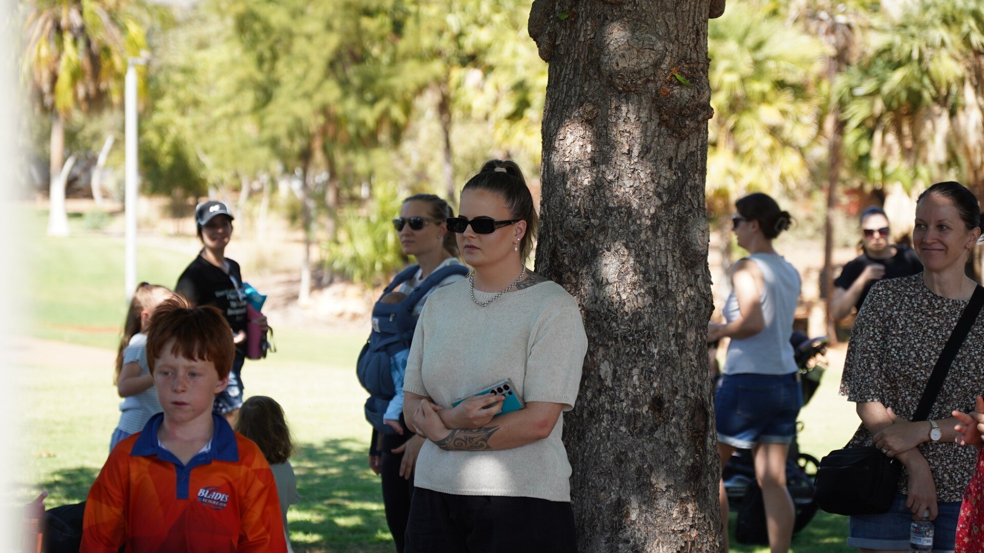 Una mujer con una blusa color crema se encuentra frente a un árbol entre una pequeña multitud de personas. 