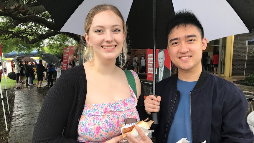 A young lady and man holding umbrella smile while holding sausage sizzle.