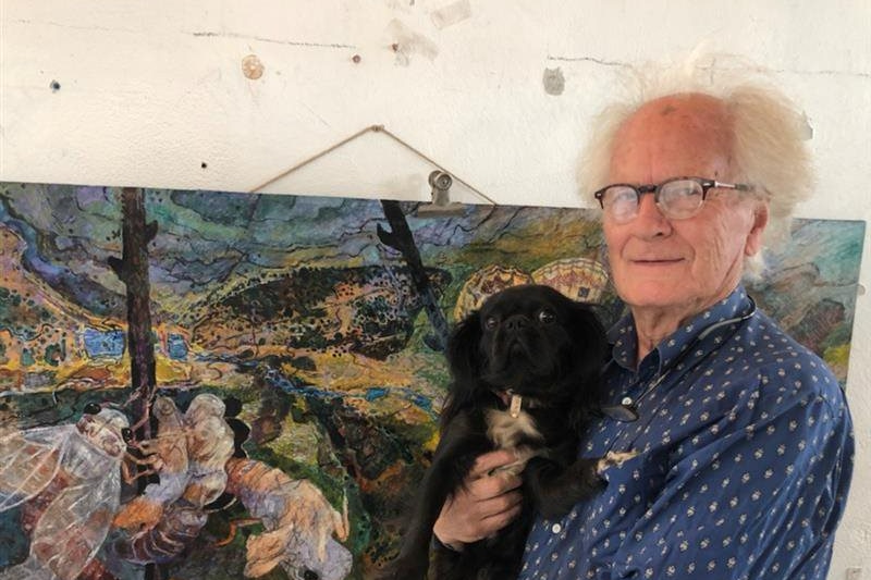 A man in glasses holds a small black dog, standing in front of a landscape artwork
