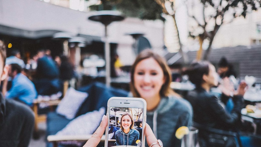 A woman at a cafe has her photo taken by a friend using a smartphone.