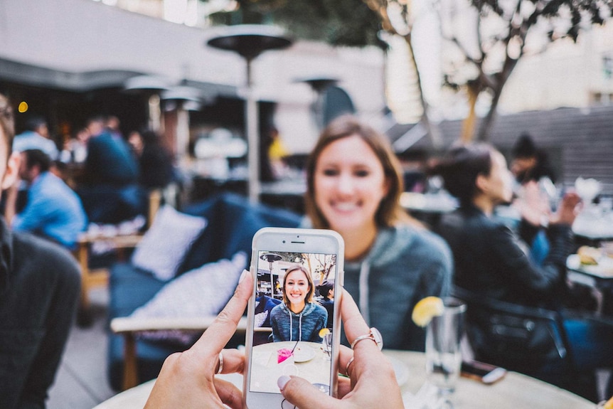 A woman at a cafe has her photo taken by a friend using a smartphone.