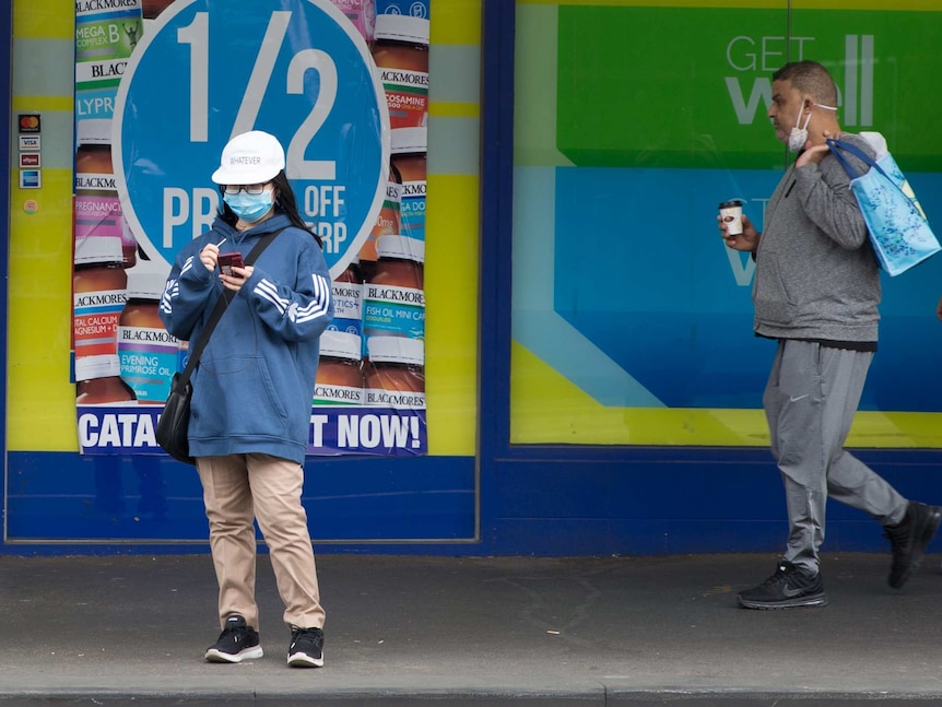 A woman wearing a mask looks at her phone in front of Chemist Warehouse as two men walk past