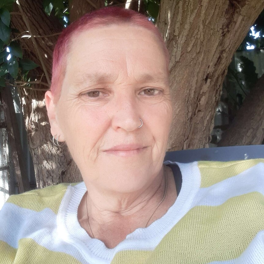 middle aged woman with shaved pink hair looks at camera.