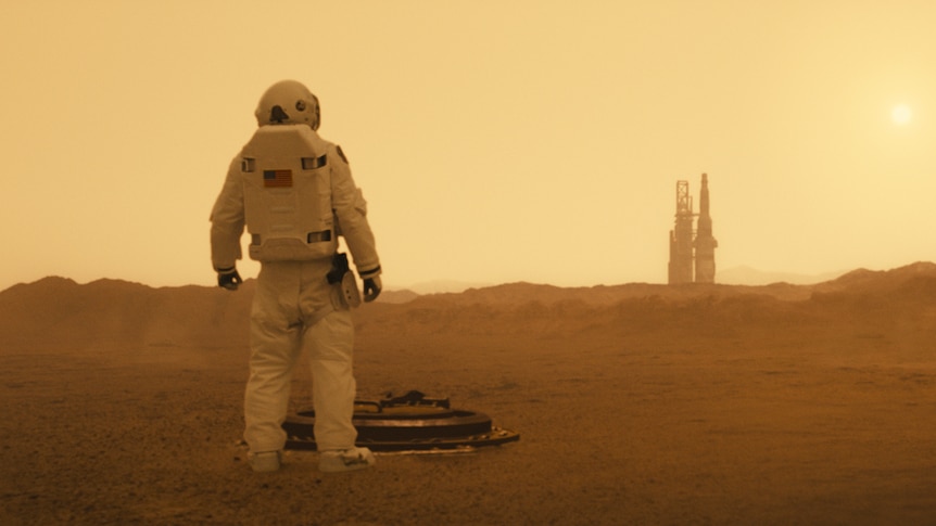 Alien landscape at sunset, with figure in midground, back turned to camera, and launch structure in distance on horizon.