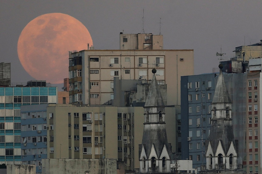 An orange coloured moon behind a bunch of buildings.