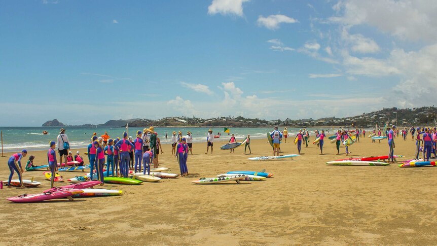 Wide photo of dozens of surf lifesavers standing on a beach with their boards and other equipment.