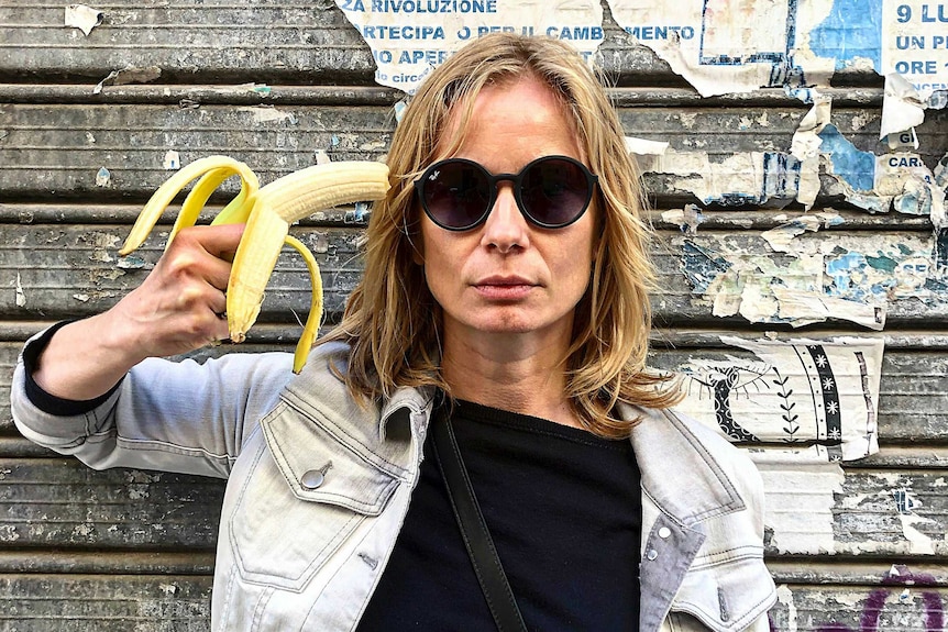 A woman in sunglasses holds a banana to her head like a gun.