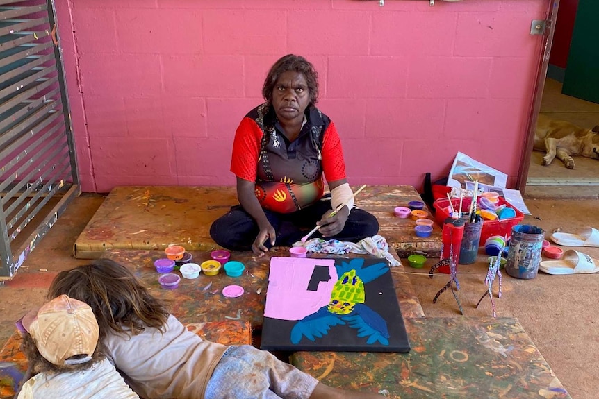 Woman sitting on ground in an art centre painting with two children lying in front of her