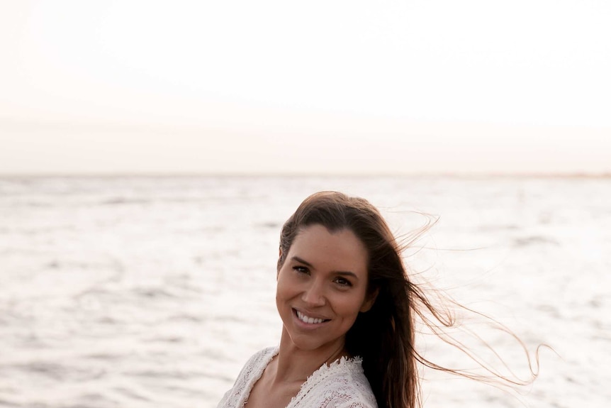 A woman with brown hair and wearing a long white dress smiles as waves crash on the sand behind her.