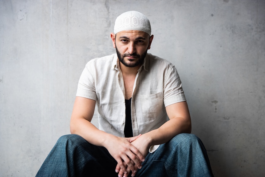 Young man with short-cropped beard and moustache wearing cream skullcap and short-sleeved white shirt, sitting with arms resting