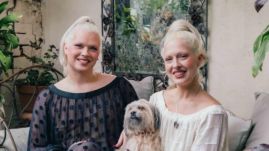 Two white women dressed in sheer dresses - one white, one black - and with matching platinum blonde hair sit smiling.
