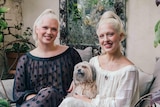 Two white women dressed in sheer dresses - one white, one black - and with matching platinum blonde hair sit smiling.