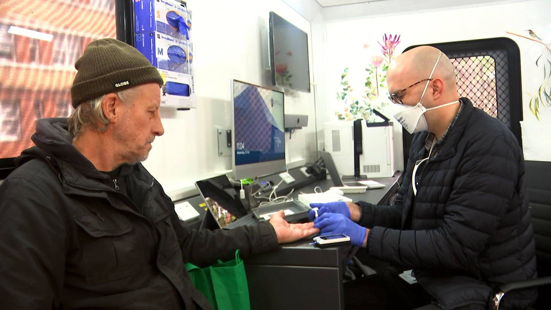 A man in a green beanie and hoodie sits across from a man in a mask, gloves and a puffer jacket inside the mobile clinic