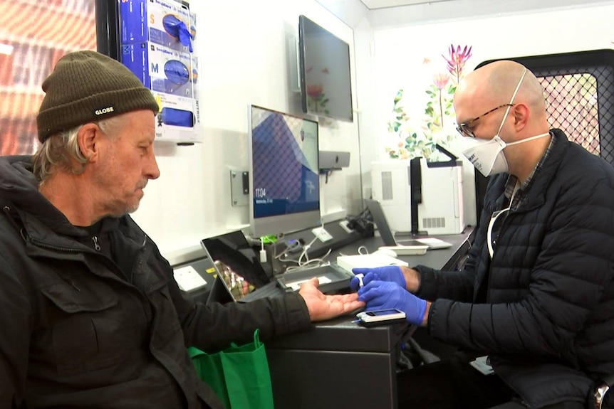 A man in a green beanie and hoodie sits across from a man in a mask, gloves and a puffer jacket inside the mobile clinic