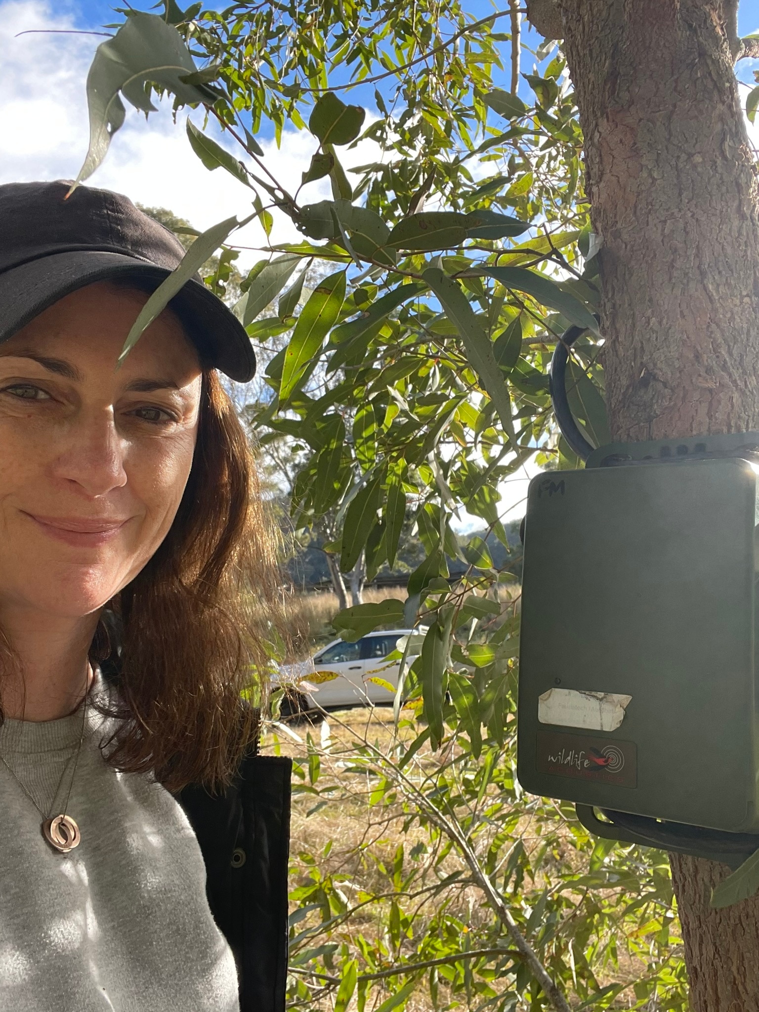 A woman with brown hair, wearing a cap, stands outdoors, with a gum tree in the background.