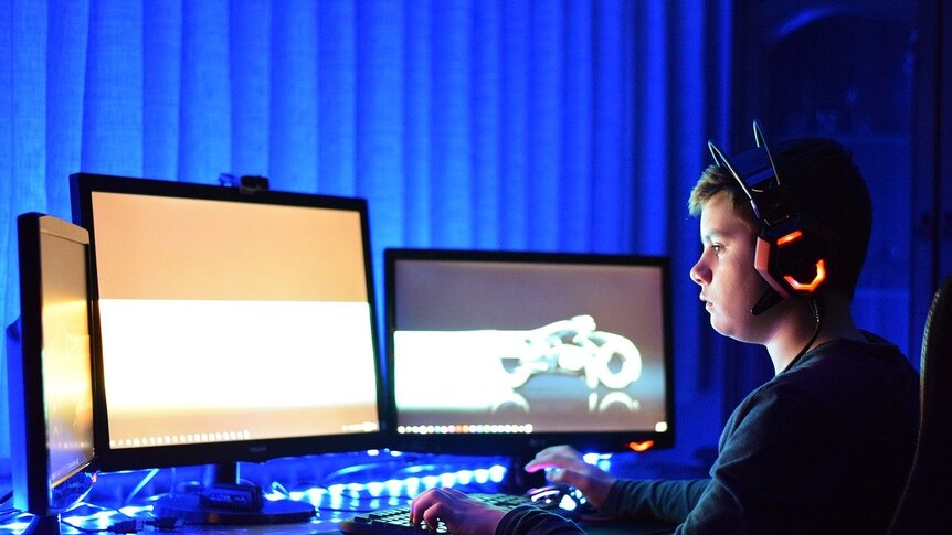 A boy in front of a computer with three screens in a dark room.