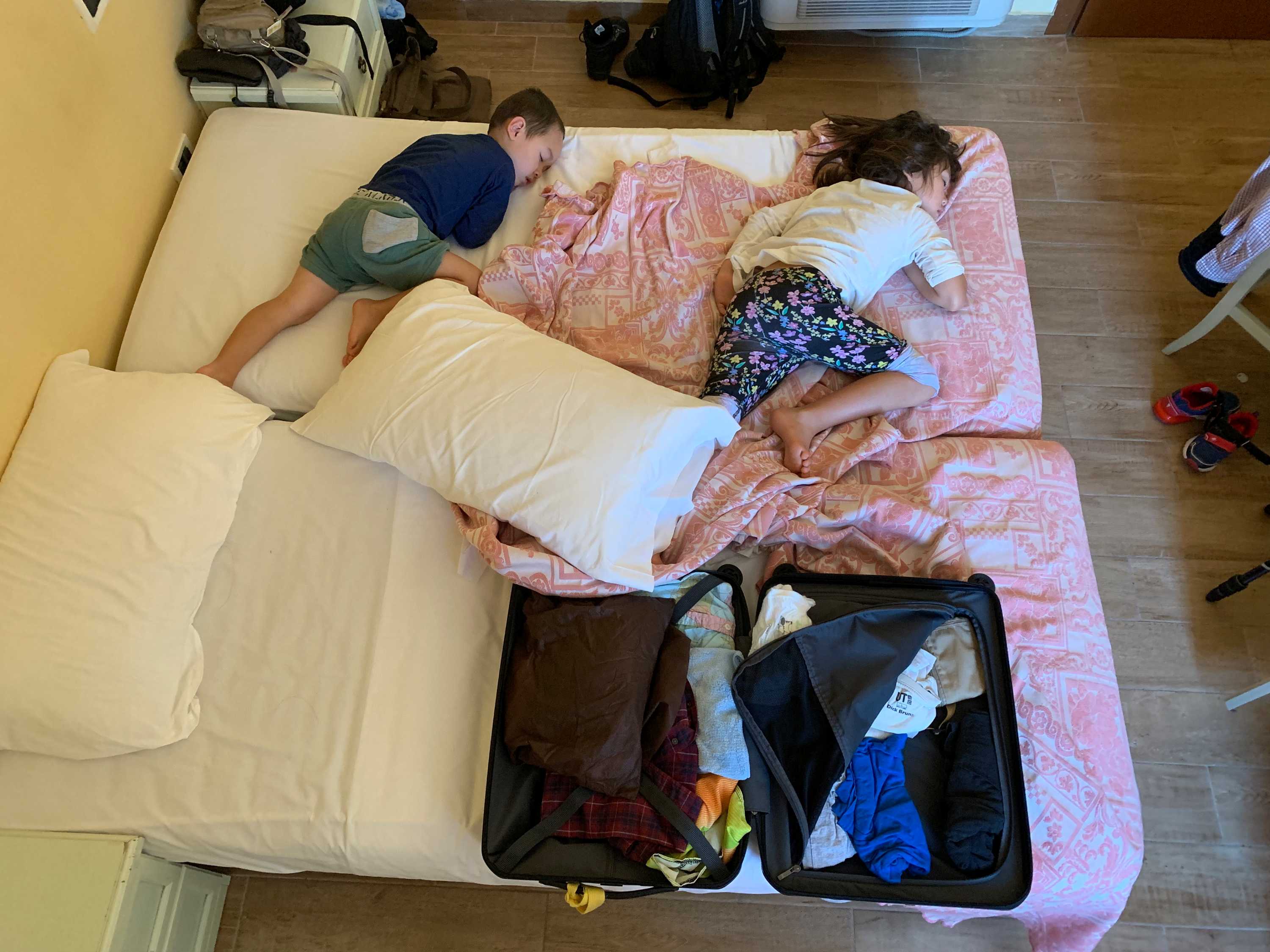 A small boy and girl sleep sideways on a bed, next to an opened and unpacked suitcase