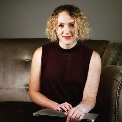 Emily Atkin sitting on a couch, she has short curly hair and is holding a laptop.