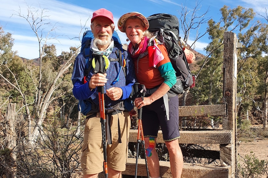 A man and a woman in hiking hear standing in front of a fence, smiling at the camera