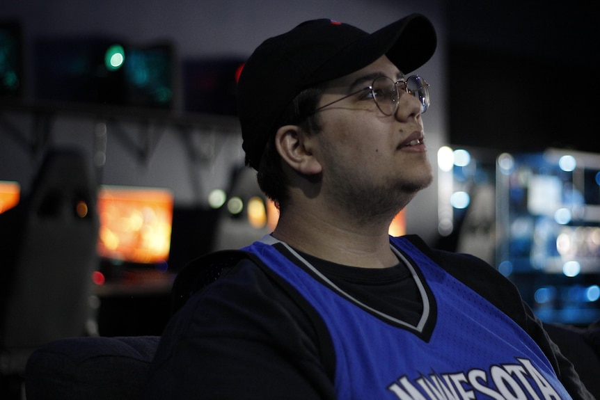 A young man with glasses and a black cap, wearing a black shirt and blue baskeball jersey, looks to his left.