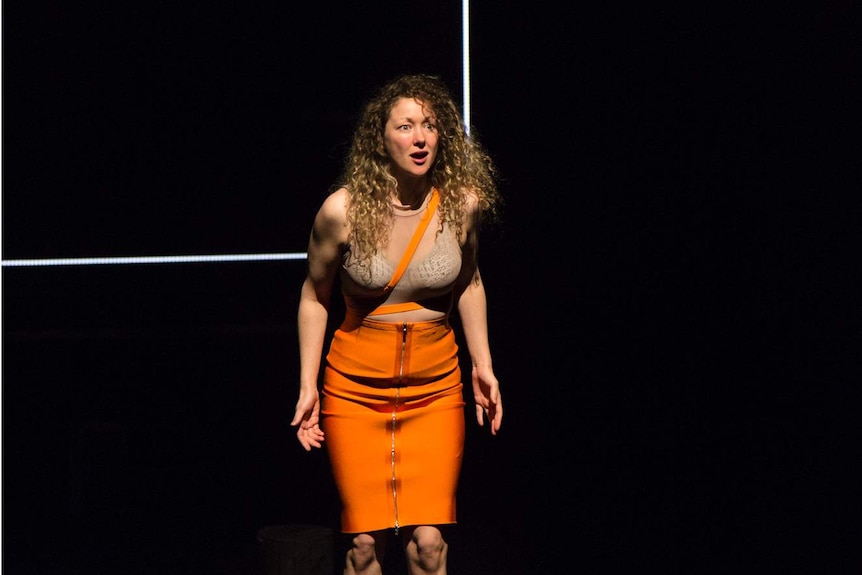 A woman on stage stands on a log, she looks angry/defiant