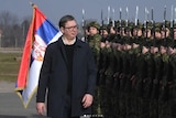 A man in a suit walks past a line of soldiers, a Serbian flag waves behind him.