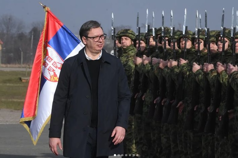 A man in a suit walks past a line of soldiers, a Serbian flag waves behind him.