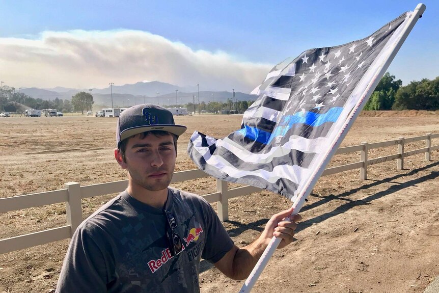 Brandon Apelian holds a black and white american flag as smoke rises in the background