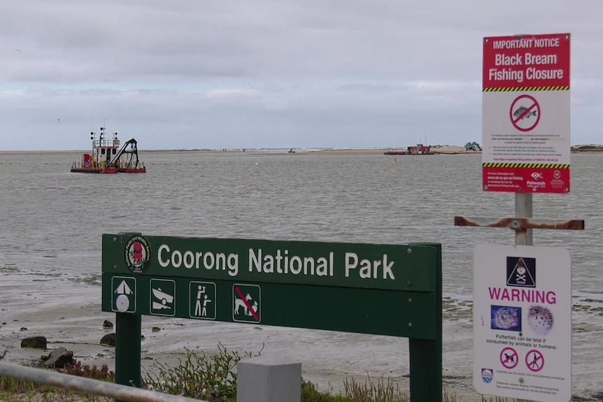 A sign saying Coorong National Park with a river, a dredging boat and the ocean behind