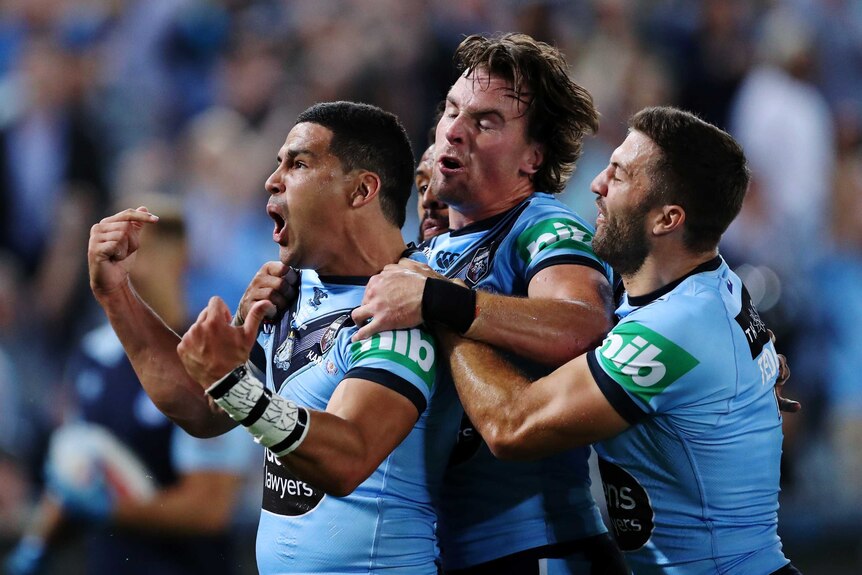A male New South Wales State of Origin player pumps both his fists as he embraced by his teammates.