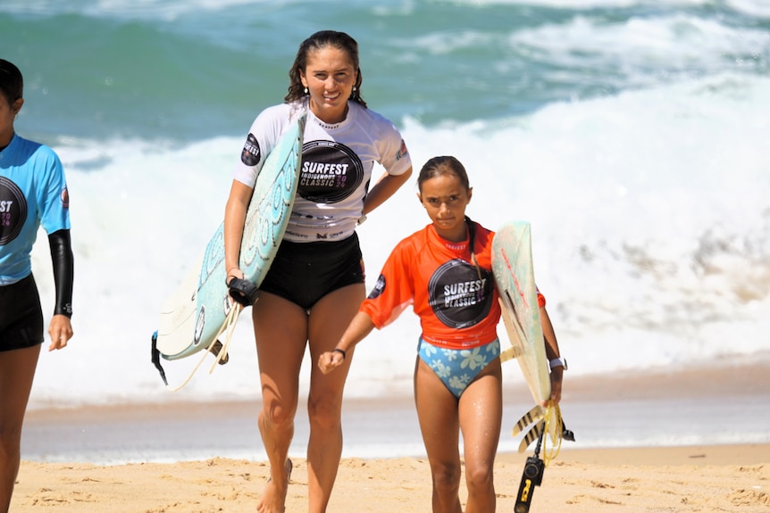 Two young women hold surfboards as they walk away from the ocean's edge.