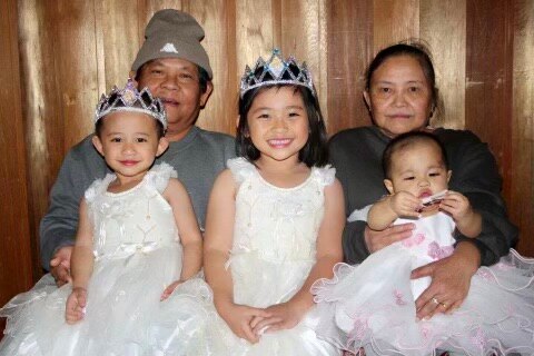 A happy family of two  grandparents with three little girls wearing paper crowns and bright white dresses on their laps.
