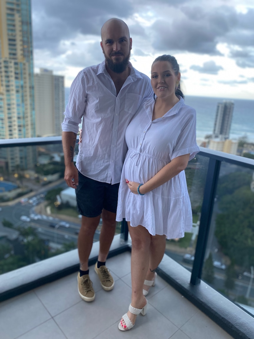 A pregnant woman stands beside her husband on an apartment balcony, as they smile to the camera.