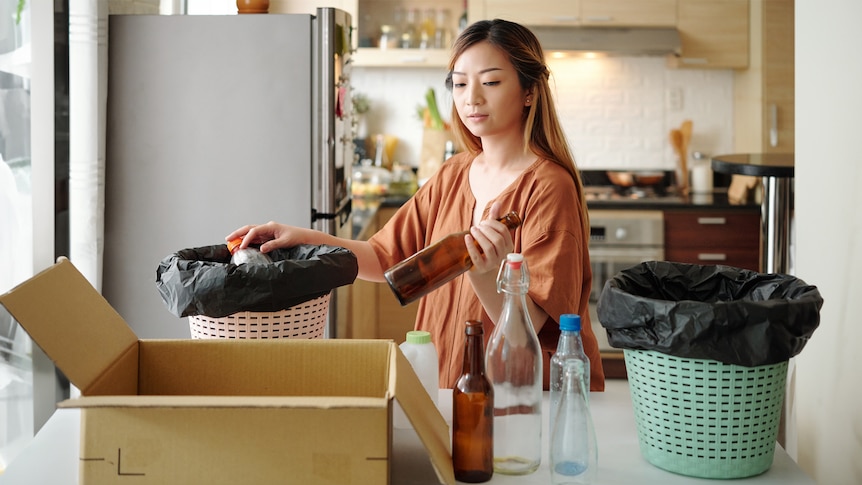 A woman hold glass bottles and sorts recycling into different tubs on a kitchen bench.