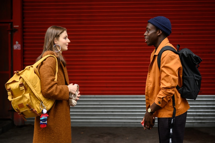A young white woman stands talking to a young Black man in front of a roller door
